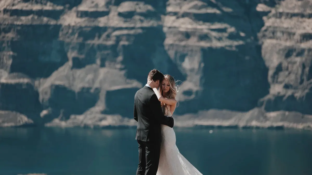A couple in wedding clothes hugging by a lake below tall cliffs