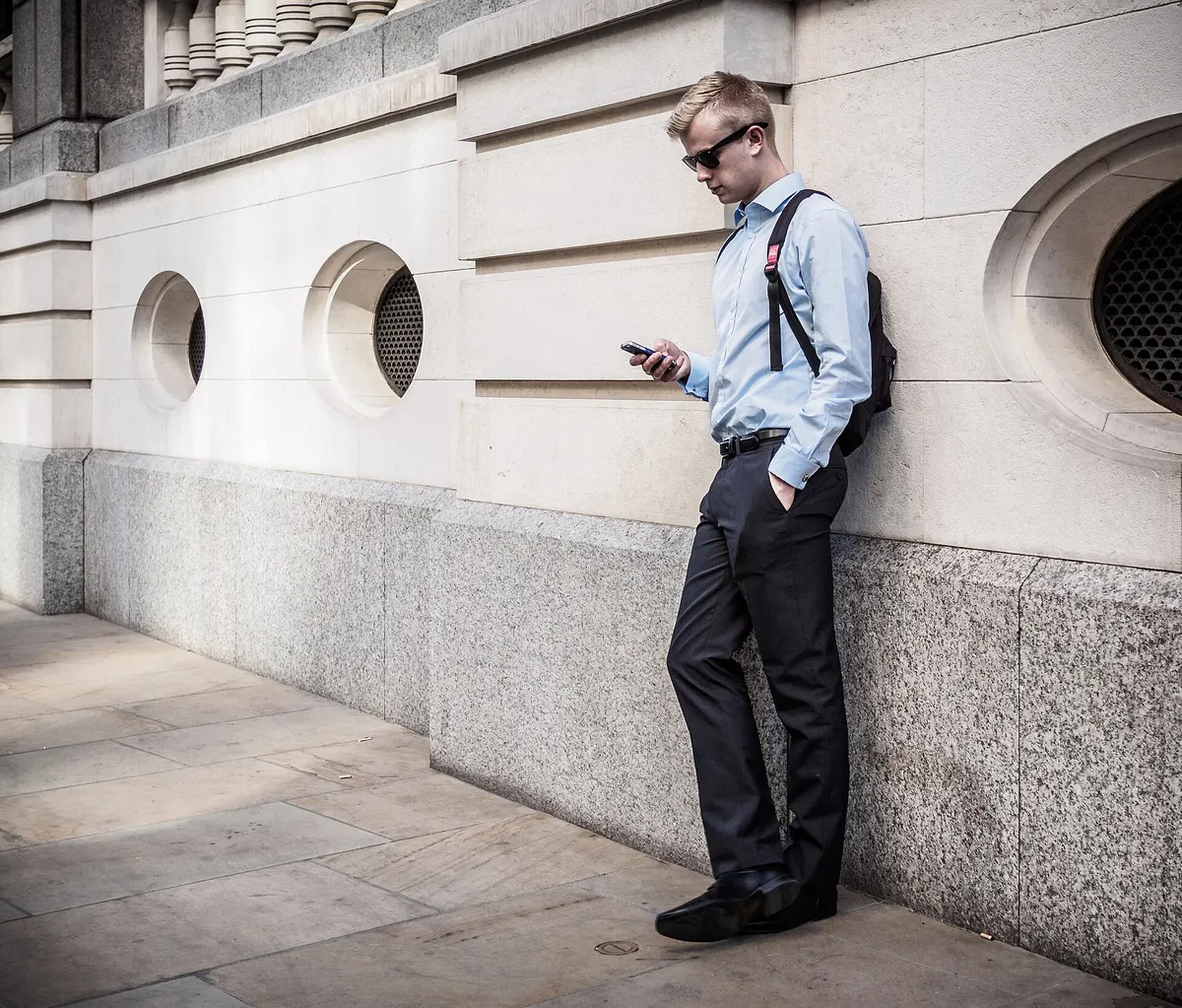 Young man in sunglasses leaning on a wall and checking his phone