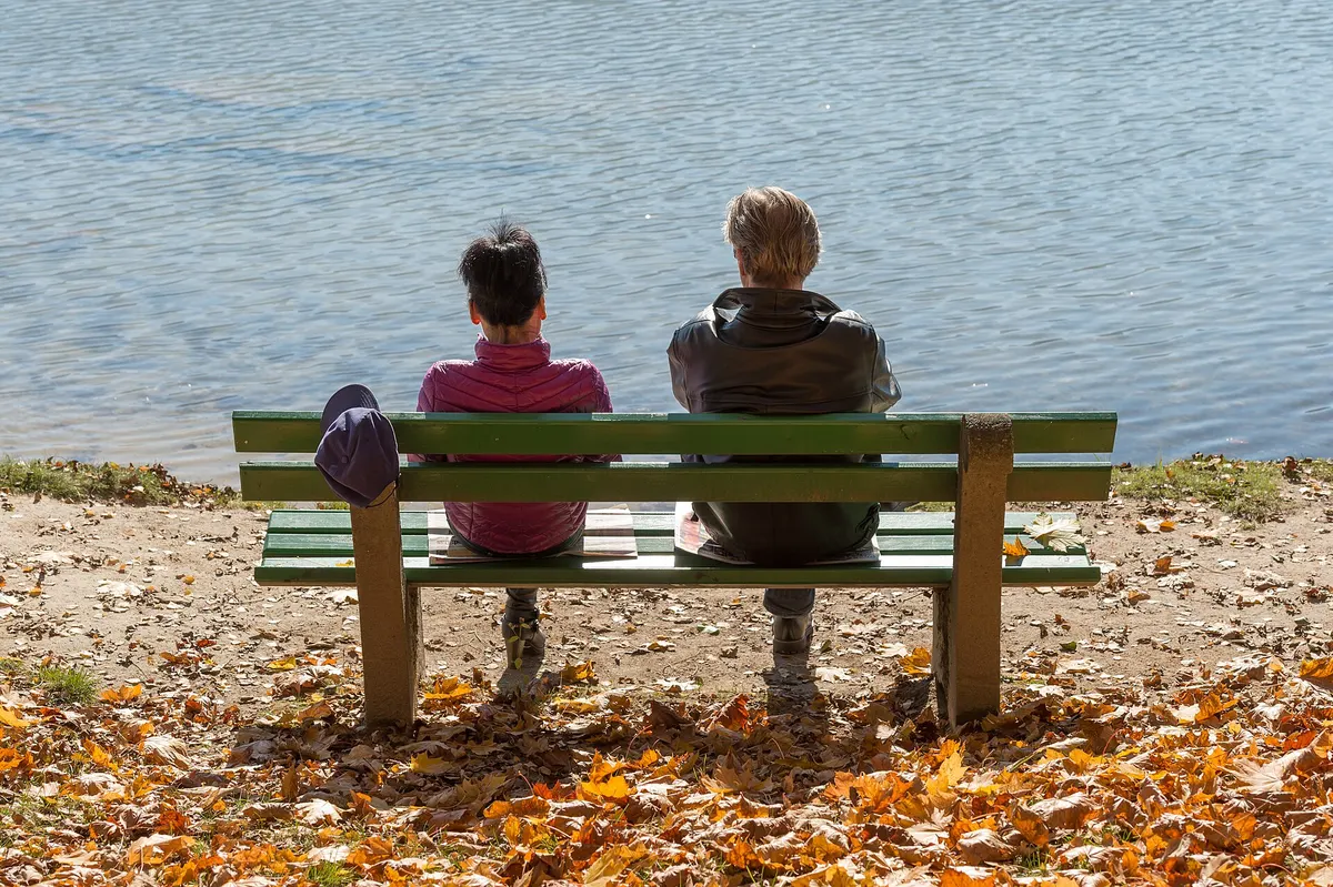 Couple on a lakeside bench among autumn leaves, seen from behind