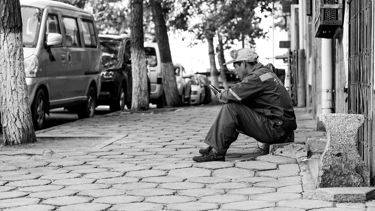 Man sitting on a cobblestone curb texting on his phone, black and white